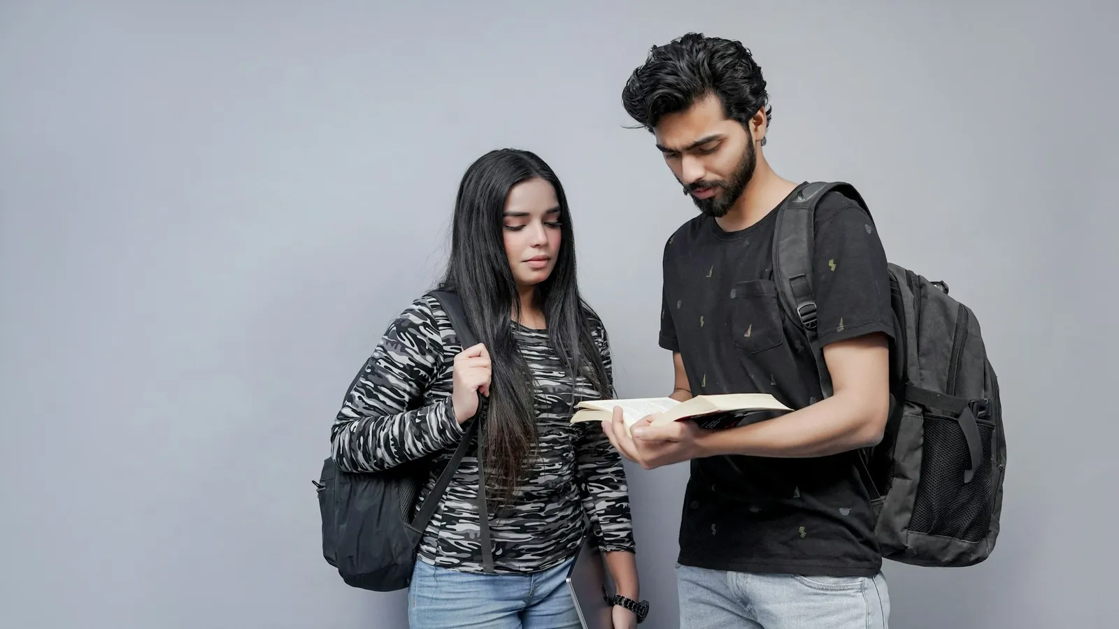 Two students with backpacks looking at a book together