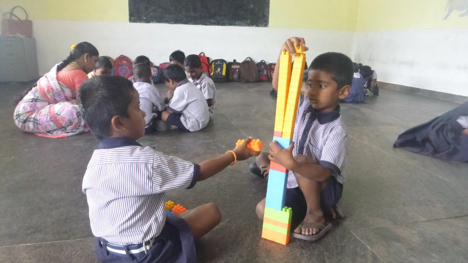 Two young children in school uniforms build a tall block tower on a classroom floor