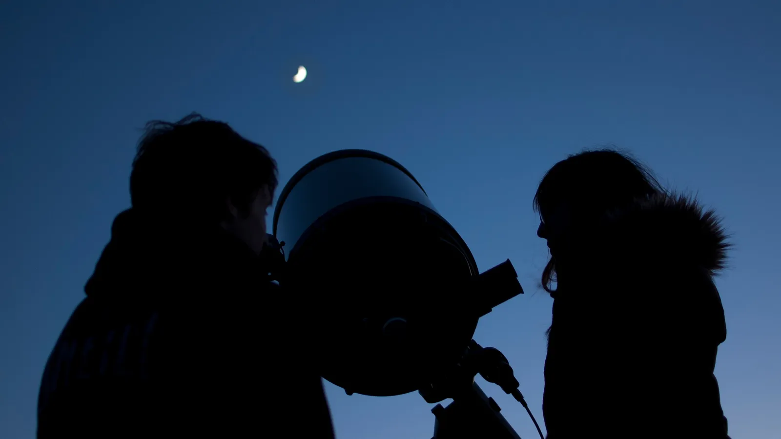 Two young people in silhouette beside a telescope under a crescent Moon