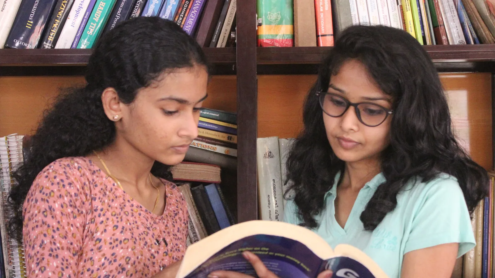 Two young women in Kerala reading a textbook together in front of a bookshelf