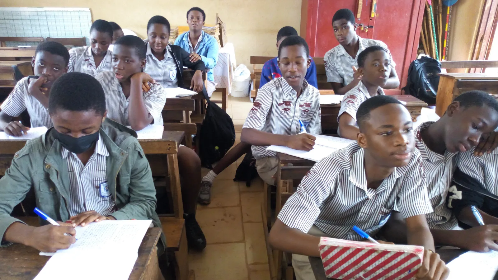 Uniformed secondary school students at wooden desks, some writing notes in exercise books