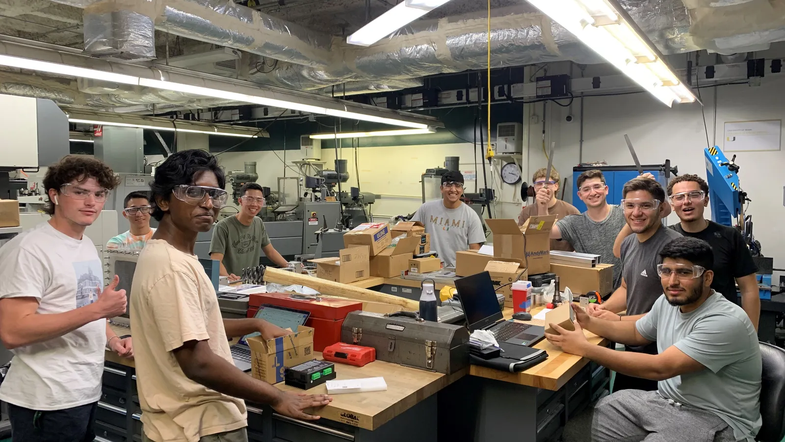 University robotics team in safety glasses gathered around workshop benches with laptops and parts
