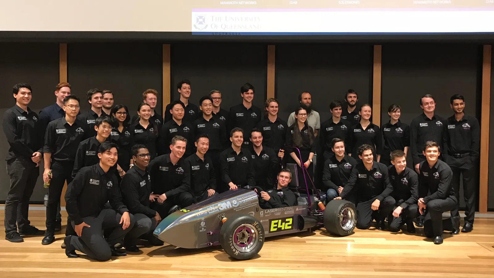University student racing team in matching black shirts posing around their single-seat race car