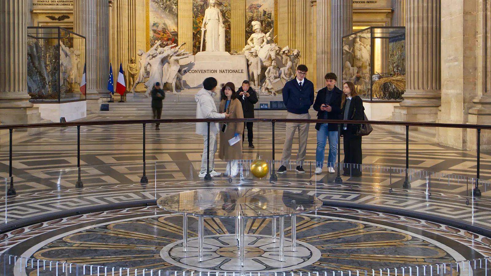 Visitors watching a long pendulum swing over a circular floor in a domed hall