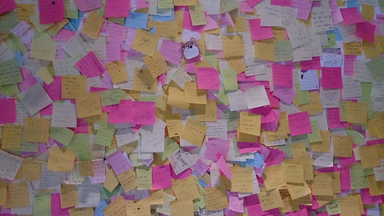 Wall covered edge to edge in overlapping handwritten sticky notes in pink, yellow, green and blue