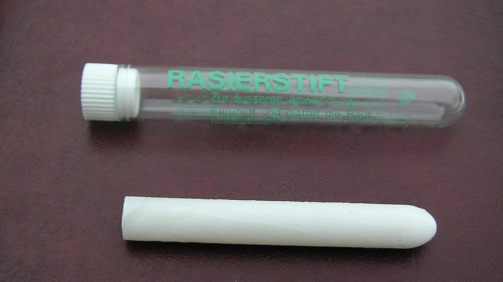 White styptic pencil lying beside its clear plastic tube on a maroon surface