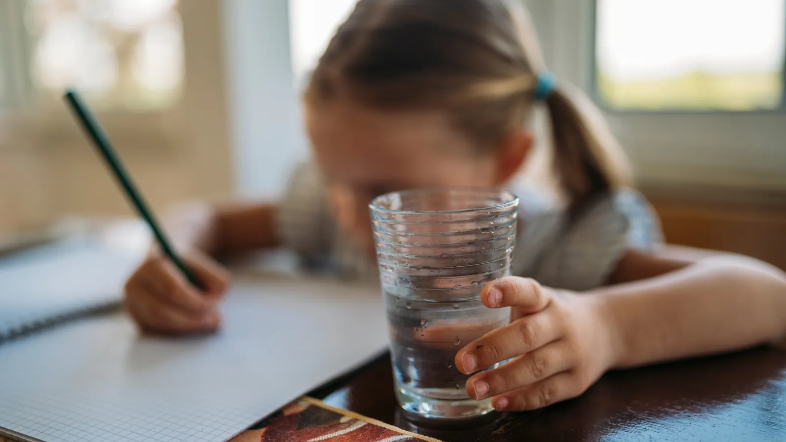 Young child bent over a squared notebook, pencil in hand, holding a glass