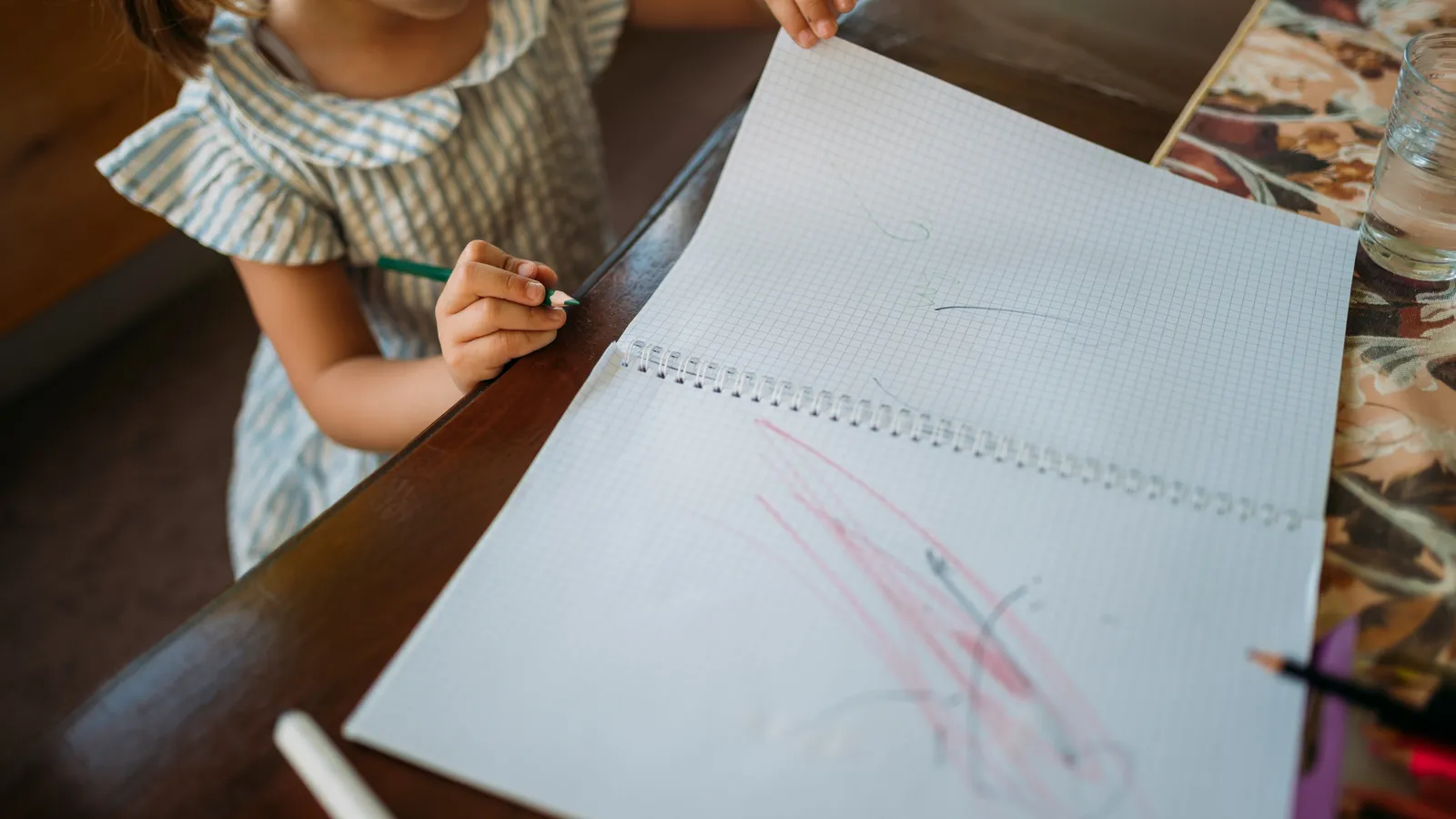 Young child holding a green pencil and drawing in a squared-paper notebook