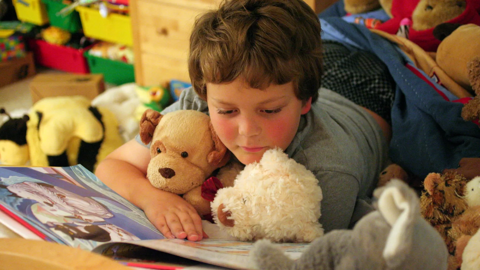 Young child lying on the floor reading a comic, hugging soft toys