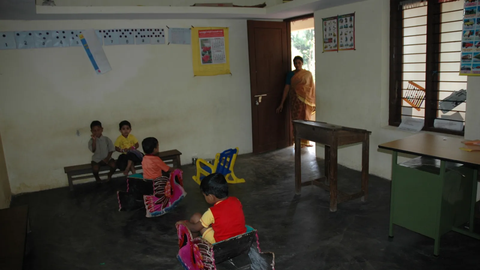Young children on a bench and rocking toys in an anganwadi classroom, a worker at the door