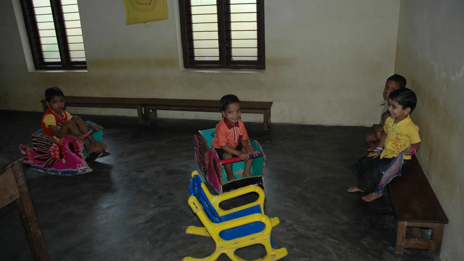 Young children on rocking toys and a bench in a small Anganwadi classroom