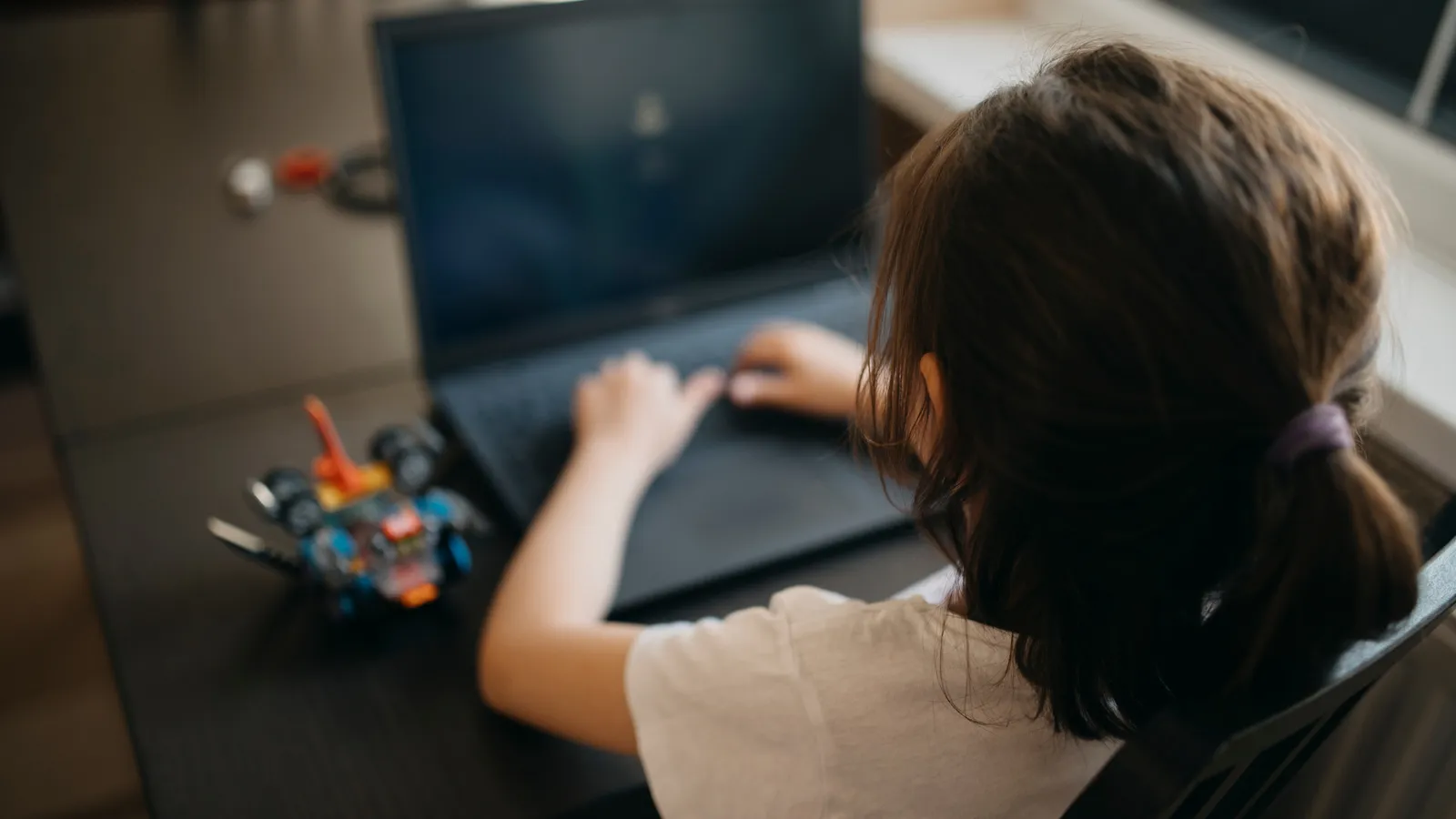 Young learner typing on a laptop at home beside a small robot car kit