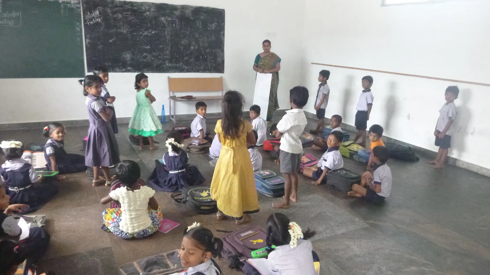 Young primary pupils with school bags sitting and standing on a classroom floor as their teacher looks on
