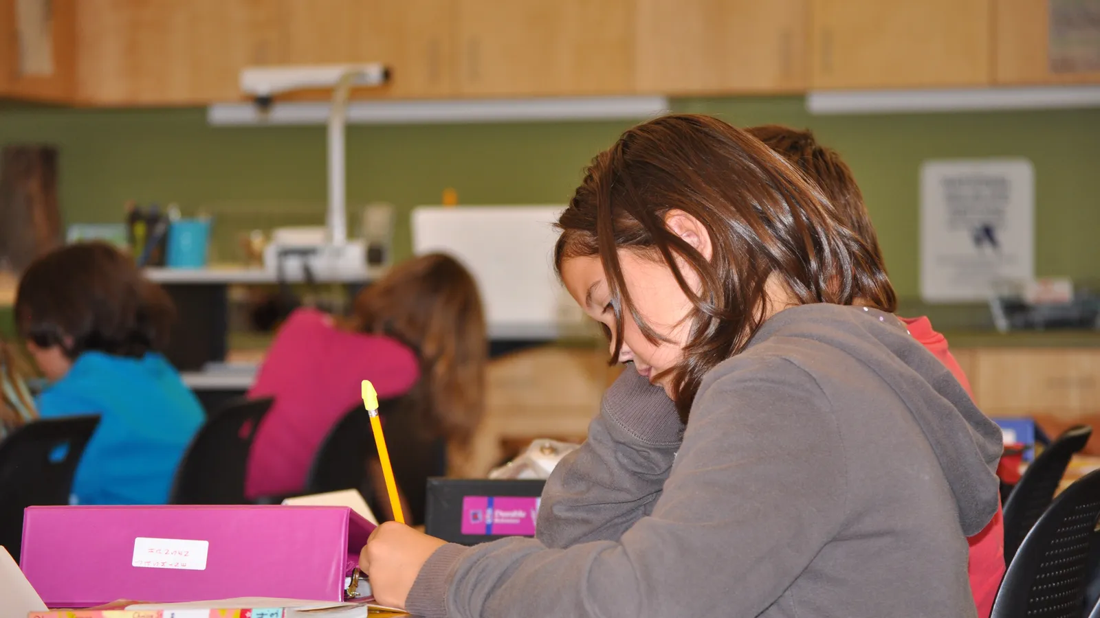 Young student bent over a classroom desk writing with a yellow pencil